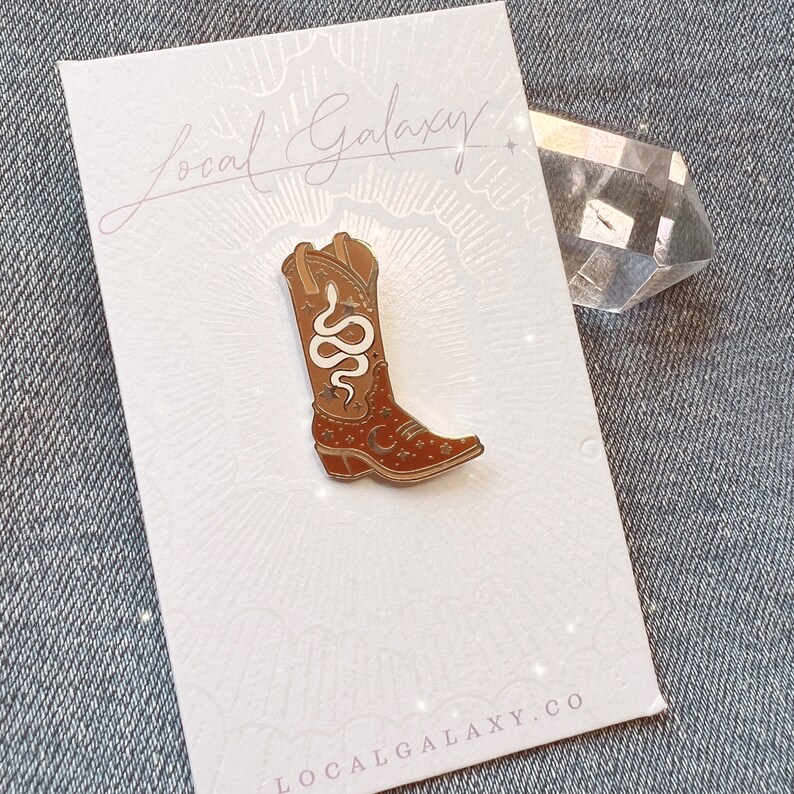 Op de afbeelding: Een gouden emaille pin met een bruine cowboylaars met een wit slang- en sterrenontwerp. De pin is op een witte kaart met de tekst "Local Galaxy" en "localgalaxy.co".