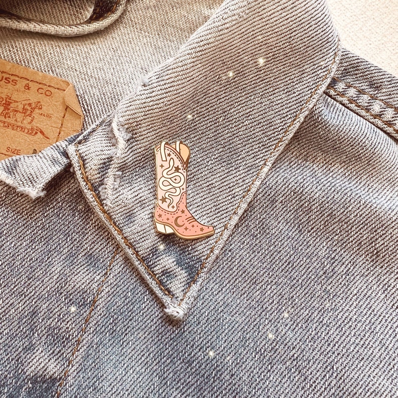 Op de afbeelding: Een roze cowboylaars emaille pin met een slang en maan ontwerp. De pin is op een spijkerjack.
