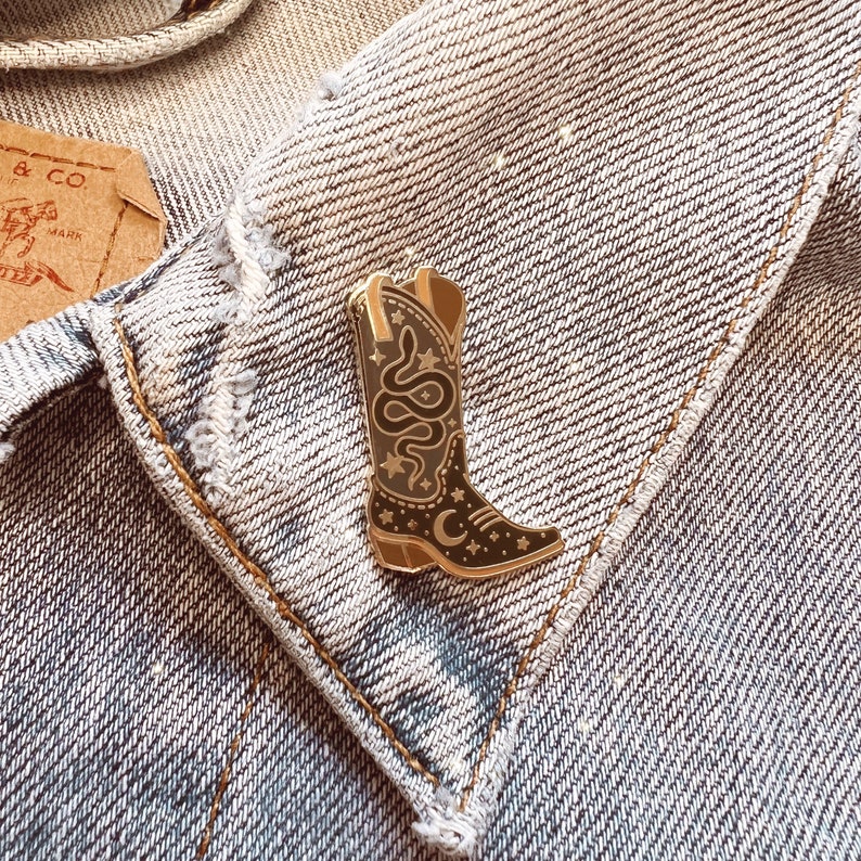 Op de afbeelding: Gouden emaille pin met een zwarte cowboylaars met een slang en halve maan ontwerp.