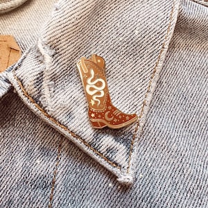 Op de afbeelding: Een gouden emaille pin met een bruine cowboylaars met een witte slang, sterren en een halve maan.