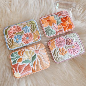 May include: Four rectangular embroidered patches with floral designs. Each patch features a different color scheme, including blue, orange, pink, and yellow flowers with green leaves. The patches are bordered with coordinating colors.