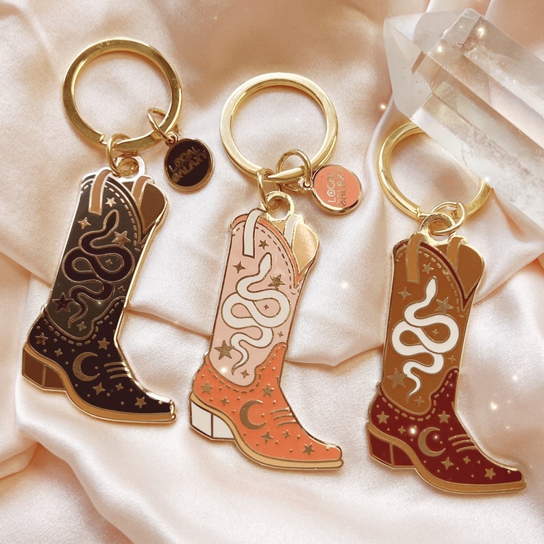May include: Three gold-toned keychains with cowboy boot charms in black, pink, and brown. Each boot features a snake design, stars, and a crescent moon. The keychains have a small circular charm that reads "Local Galaxy".