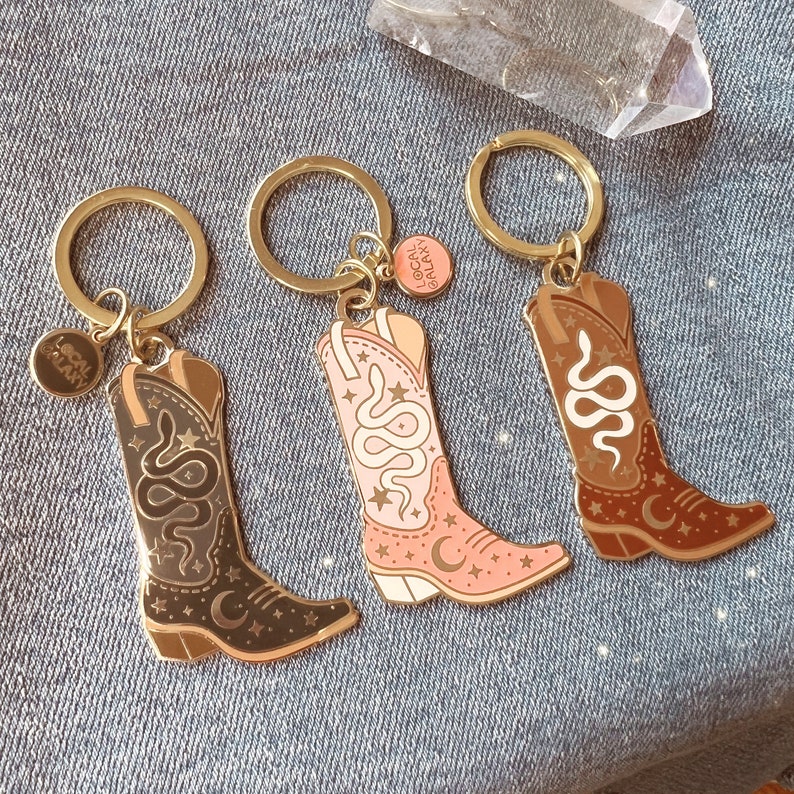 May include: Three keychains with gold-colored rings and charms. Each keychain features a cowboy boot design with a snake and celestial details. The boots are in black, pink, and brown, with the text "Local Galaxy" on the charm.