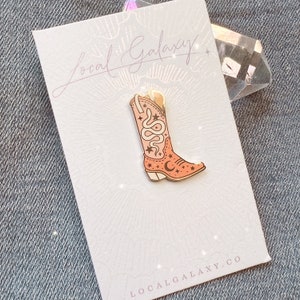Op de afbeelding: Een roze emaille pin met een cowboylaars met een slang- en sterrenontwerp. De pin is op een witte kaart met de tekst "Local Galaxy" en "localgalaxy.co".