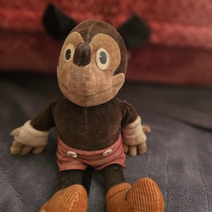 Op de afbeelding: Vintage pluche speelgoed van een stripfiguur met een bruin lichaam, een beige gezicht en grote witte ogen. Het speelgoed draagt een roze broek en oranje schoenen. Zwarte oren zijn zichtbaar achter het hoofd. Het speelgoed staat op een blauw oppervlak.