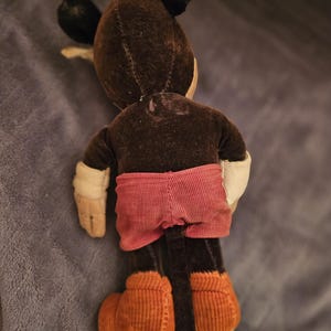 Op de afbeelding: Vintage Mickey Mouse pluche speelgoed, van achteren gezien. Het pluche heeft een bruin lichaam, zwarte oren en een rode corduroy broek. De handen zijn wit en de schoenen zijn van oranje corduroy.