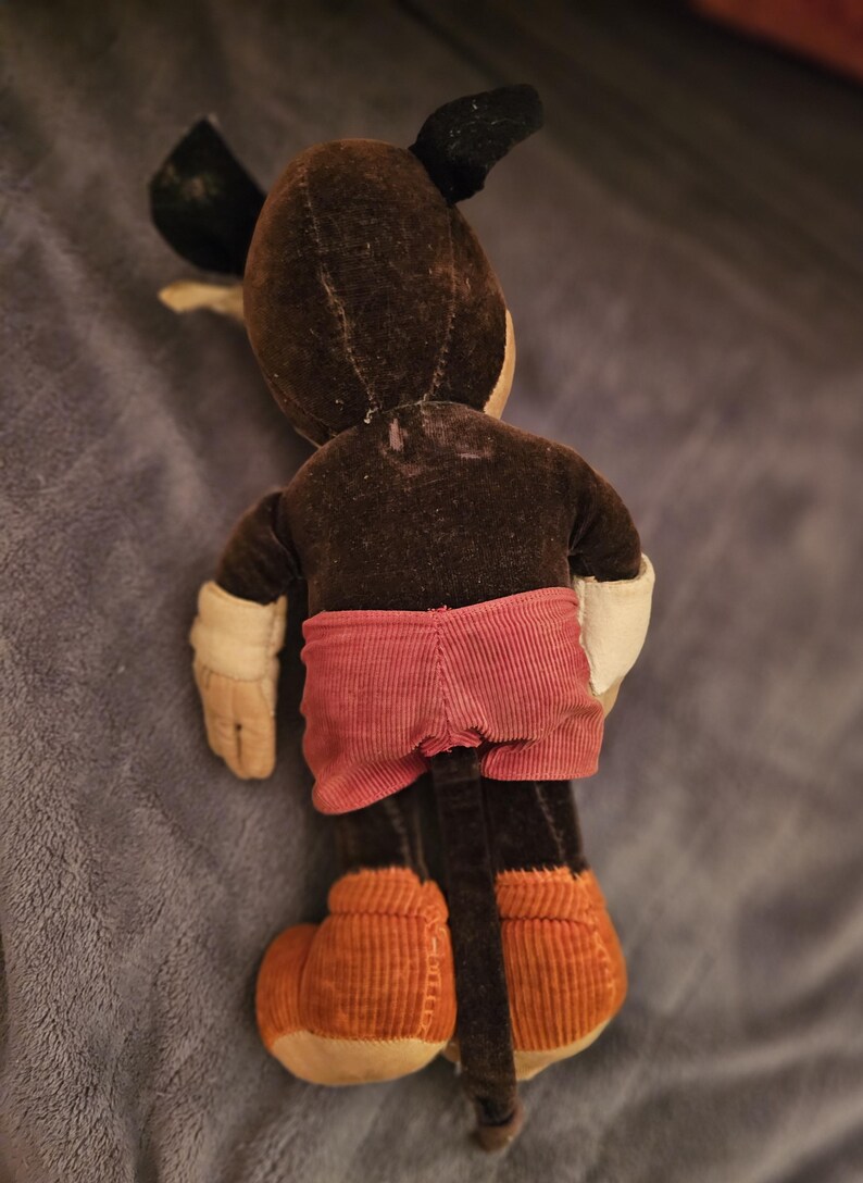 Op de afbeelding: Vintage Mickey Mouse pluche speelgoed, van achteren gezien. Het pluche heeft een bruin lichaam, zwarte oren en een rode corduroy broek. De handen zijn wit en de schoenen zijn van oranje corduroy.