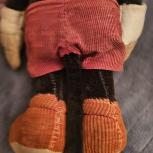 Op de afbeelding: Een pluche speelgoed met een bruin lichaam, rode corduroy shorts en oranje corduroy schoenen. Het speelgoed heeft witte handschoenen en een lange zwarte staart. Het speelgoed is een klassiek karakter.