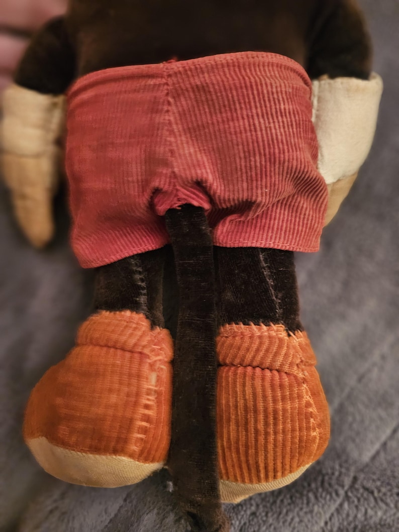 Op de afbeelding: Een pluche speelgoed met een bruin lichaam, rode corduroy shorts en oranje corduroy schoenen. Het speelgoed heeft witte handschoenen en een lange zwarte staart. Het speelgoed is een klassiek karakter.