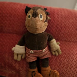 Op de afbeelding: Een vintage pluche apenpop met een bruin lichaam, gezicht en oren. Het heeft witte handschoenen, een roze korte broek met witte knopen en oranje corduroy schoenen. De pop zit op een rode, getextureerde ondergrond.