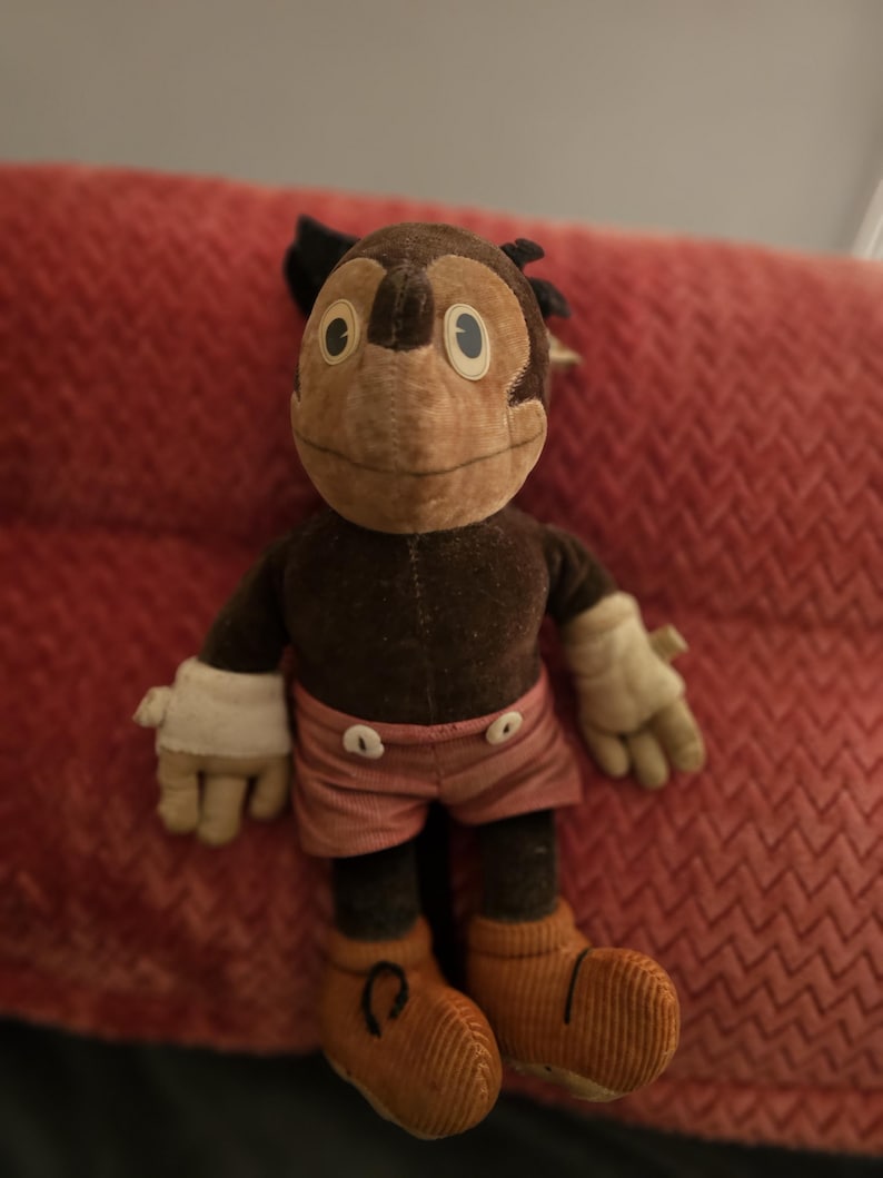 Op de afbeelding: Een vintage pluche apenpop met een bruin lichaam, gezicht en oren. Het heeft witte handschoenen, een roze korte broek met witte knopen en oranje corduroy schoenen. De pop zit op een rode, getextureerde ondergrond.