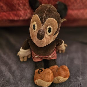 Op de afbeelding: Vintage Mickey Mouse pluche pop. De pop heeft een bruine kop en lichaam, met grote witte ogen en een zwarte neus. Het draagt een roze broek, witte handschoenen en oranje schoenen. De pop zit op een blauw oppervlak.