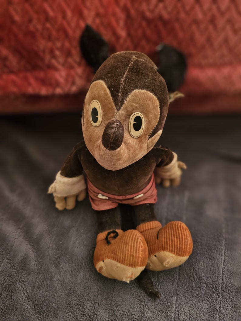 Op de afbeelding: Vintage Mickey Mouse pluche pop. De pop heeft een bruine kop en lichaam, met grote witte ogen en een zwarte neus. Het draagt een roze broek, witte handschoenen en oranje schoenen. De pop zit op een blauw oppervlak.