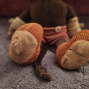 Op de afbeelding: Een vintage knuffel aap, gezien van achteren, met oranje corduroy voeten en een bruin lichaam. De aap draagt een roze en wit gestreepte korte broek. De voeten vertonen tekenen van slijtage, wat bijdraagt aan de charme.