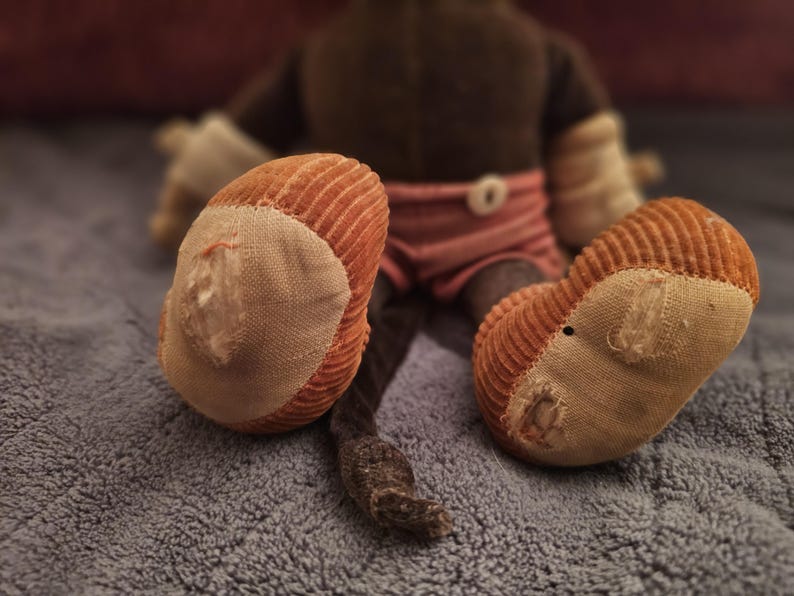 Op de afbeelding: Een vintage knuffel aap, gezien van achteren, met oranje corduroy voeten en een bruin lichaam. De aap draagt een roze en wit gestreepte korte broek. De voeten vertonen tekenen van slijtage, wat bijdraagt aan de charme.