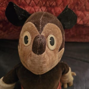 Op de afbeelding: Vintage Mickey Mouse pluche speelgoed. De pluche heeft een bruine kop en lichaam met zwarte oren, beige handen en een rode broek. De ogen zijn groot en ovaal. Het speelgoed is gemaakt van een zachte, versleten stof, wat suggereert dat het een antiek verzamelobject is.
