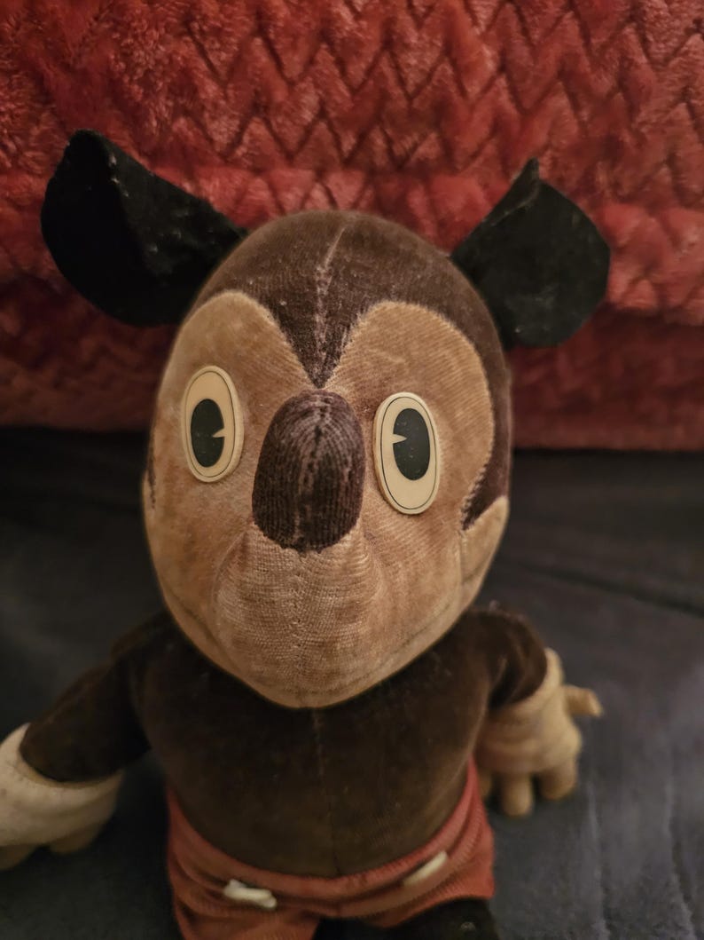 Op de afbeelding: Vintage Mickey Mouse pluche speelgoed. De pluche heeft een bruine kop en lichaam met zwarte oren, beige handen en een rode broek. De ogen zijn groot en ovaal. Het speelgoed is gemaakt van een zachte, versleten stof, wat suggereert dat het een antiek verzamelobject is.