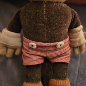 Op de afbeelding: Een vintage aap knuffel, gezien van achteren. De aap heeft een bruin lichaam, armen en benen, met roze gestreepte shorts en witte handschoenen. De shorts zijn vastgemaakt met twee witte knopen. De voeten van de aap zijn oranje.