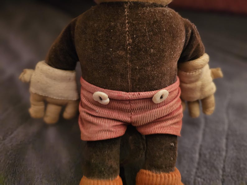 Op de afbeelding: Een vintage aap knuffel, gezien van achteren. De aap heeft een bruin lichaam, armen en benen, met roze gestreepte shorts en witte handschoenen. De shorts zijn vastgemaakt met twee witte knopen. De voeten van de aap zijn oranje.