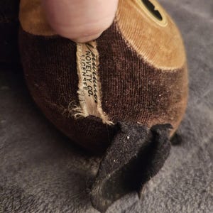 Op de afbeelding: Close-up van een vintage knuffel, voornamelijk bruin en bruin met een zwarte oor. Het label luidt "American Novelty Co. Licensee". Het pluche speelgoed heeft een cirkelvormig oogdetail en is gemaakt van een zachte, getextureerde stof.