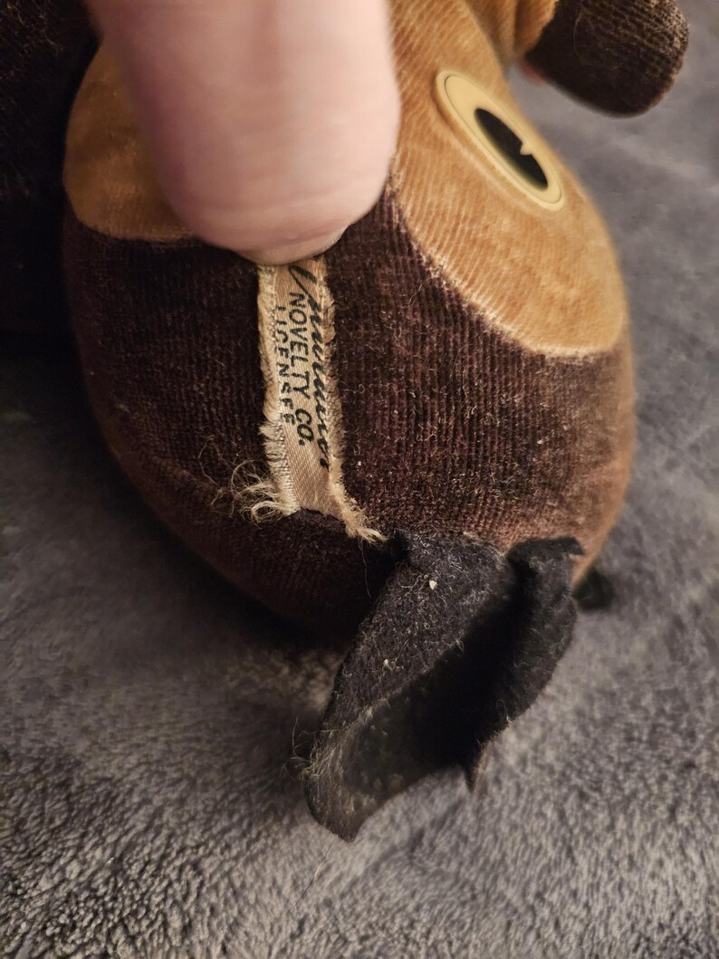 Op de afbeelding: Close-up van een vintage knuffel, voornamelijk bruin en bruin met een zwarte oor. Het label luidt "American Novelty Co. Licensee". Het pluche speelgoed heeft een cirkelvormig oogdetail en is gemaakt van een zachte, getextureerde stof.