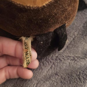 Op de afbeelding: Close-up van een vintage pluche speelgoed, met bruine en bruine stof met een zwart accent. Een klein, versleten label leest "Mickey Mouse". Het speelgoed ligt op een grijze, getextureerde ondergrond.