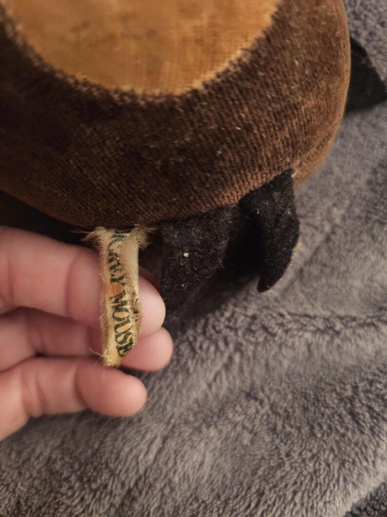 Op de afbeelding: Close-up van een vintage pluche speelgoed, met bruine en bruine stof met een zwart accent. Een klein, versleten label leest "Mickey Mouse". Het speelgoed ligt op een grijze, getextureerde ondergrond.