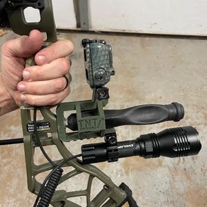 May include: An olive green compound bow with a black flashlight and a camera mount. The bow has a black handle and a camouflage pattern on the limbs. The flashlight is labeled "CYCLOP". The camera mount is labeled "TNT Hunting".