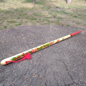 以下が含まれることがあります： 赤い房と花柄が描かれた木製の棒。棒は木の切り株の上に置かれている。