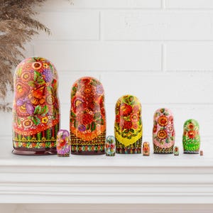 Puede incluir: Un conjunto de mu&ntilde;ecas rusas pintadas con dise&ntilde;os florales en rojo, naranja, amarillo y morado. Las mu&ntilde;ecas, de diferentes tama&ntilde;os, est&aacute;n dispuestas sobre una superficie blanca. La m&aacute;s grande mide unos 18 cm de alto.