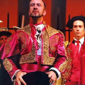 May include: Two men wearing traditional red and gold bullfighting costumes. The man in the foreground is looking up and holding a black hat in his hands. The man in the background is looking at the camera.