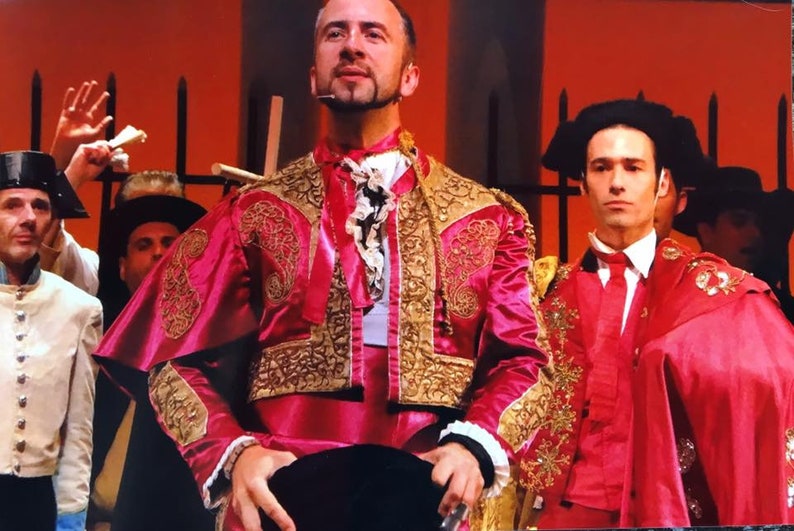 May include: Two men wearing traditional red and gold bullfighting costumes. The man in the foreground is looking up and holding a black hat in his hands. The man in the background is looking at the camera.