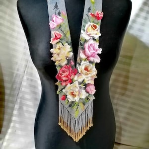 May include: A beaded necklace with a floral design. The necklace features a grey base with a pattern of pink, red, and white roses. The bottom of the necklace has a fringe of gold and white beads. The necklace is displayed on a black mannequin.