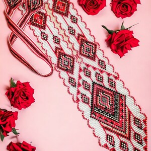 Pode incluir: Um colar de contas brancas e vermelhas com um padrão geométrico. O colar está sobre um fundo rosa com rosas vermelhas.