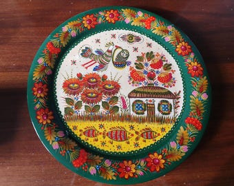 May include: A round wooden plate with a green background and a colorful folk art design. The design features a bird, a house, flowers, and fish. The plate is perfect for serving snacks or appetizers.