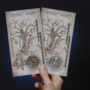 Puede incluir: Dos monedas conmemorativas en estuches protectores se muestran sobre un fondo oscuro. Las monedas se colocan sobre un fondo de papel con una ilustración de árbol y caballo. El texto "NEITHER BULLET NOR SABER" es visible.