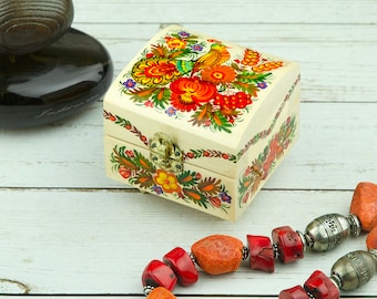 May include: A wooden box with a hinged lid, painted with colorful floral designs and a bird. The box is closed and sitting on a white wooden surface. A red coral necklace with silver accents is in the foreground.