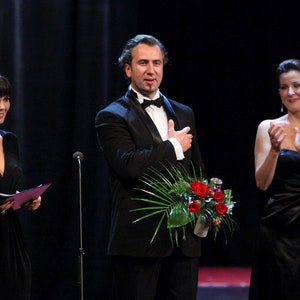 May include: Three people on stage, a man in a black tuxedo holding a bouquet of red roses, a woman in a black dress clapping, and a woman in a black dress holding a script.