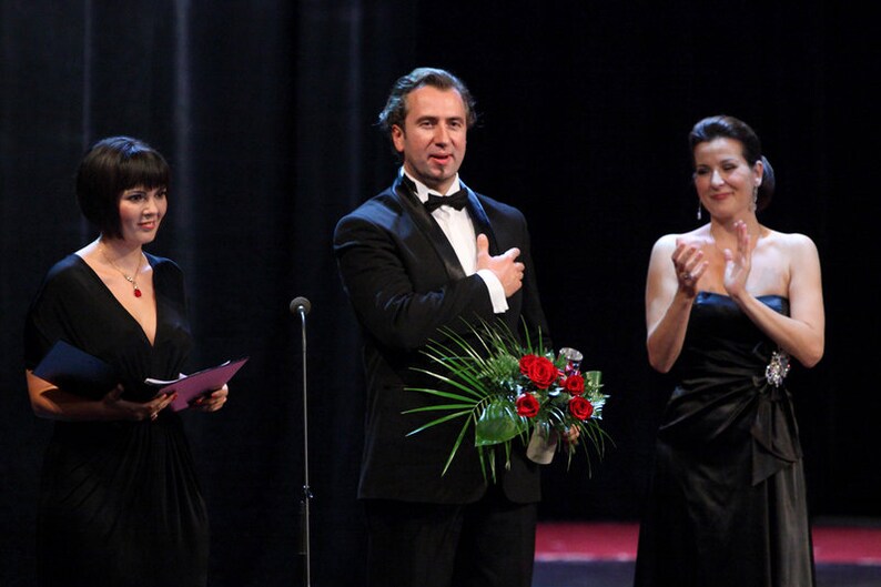 May include: Three people on stage, a man in a black tuxedo holding a bouquet of red roses, a woman in a black dress clapping, and a woman in a black dress holding a script.