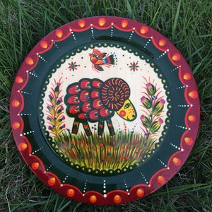 May include: A hand-painted wooden plate with a green background and a red rim. The plate features a colourful illustration of a ram with a red, orange, and yellow body, standing in a field of green grass with flowers. A small red bird with yellow accents is perched on a branch above the ram.