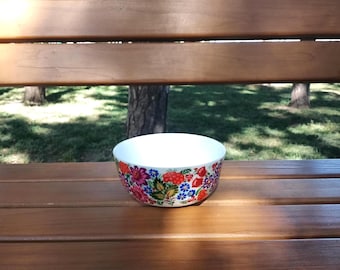 Puede incluir: Un cuenco de cerámica blanca con un vibrante patrón floral multicolor en el exterior. El diseño incluye ilustraciones detalladas de bayas rojas, acianos azules y diversas flores rosas y naranjas sobre un fondo de color blanco.
