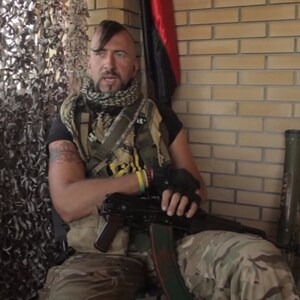 May include: A man wearing camouflage clothing and a black scarf sits holding a rifle. He has a mohawk hairstyle and a tattoo on his arm. He is sitting in front of a brick wall with a green tube and a black flag in the background.