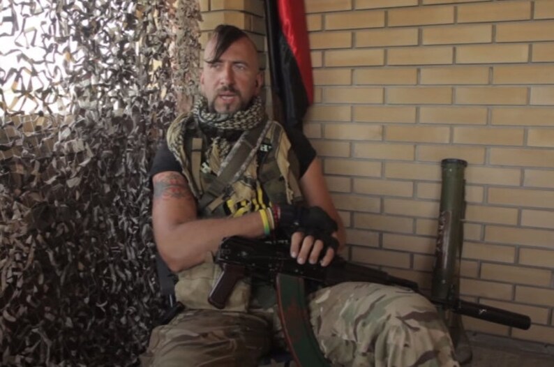 May include: A man wearing camouflage clothing and a black scarf sits holding a rifle. He has a mohawk hairstyle and a tattoo on his arm. He is sitting in front of a brick wall with a green tube and a black flag in the background.