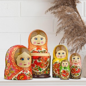 Puede incluir: Un conjunto de seis mu&ntilde;ecas rusas tradicionales, o Matrioskas, con un dise&ntilde;o floral pintado. Las mu&ntilde;ecas presentan una variedad de colores, incluyendo rojo, amarillo y verde, y est&aacute;n dispuestas sobre una superficie blanca.