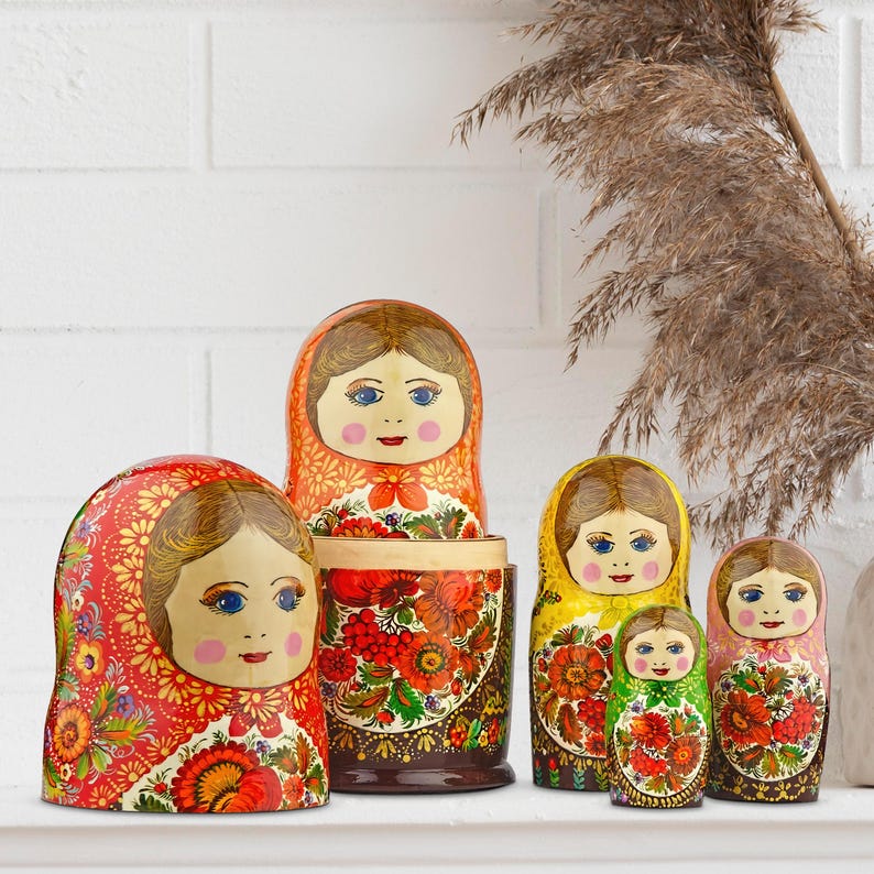 Puede incluir: Un conjunto de seis mu&ntilde;ecas rusas tradicionales, o Matrioskas, con un dise&ntilde;o floral pintado. Las mu&ntilde;ecas presentan una variedad de colores, incluyendo rojo, amarillo y verde, y est&aacute;n dispuestas sobre una superficie blanca.
