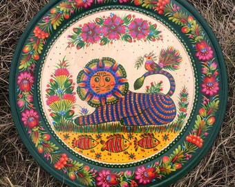 May include: A round, hand-painted wooden tray with a dark green border and a colorful floral design. The tray features a lion with a blue and orange striped body, a bird, and three fish. The background is a light beige color.
