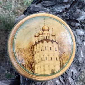 May include: A round wooden box featuring a painted image of a white church with gold domes. The church is surrounded by trees and a light brown sky. The box has a green and brown border.