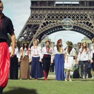 May include: A group of people wearing traditional Ukrainian clothing stand in front of the Eiffel Tower in Paris, France. The people are holding hands and smiling. A man in a black shirt and red pants walks towards the group.