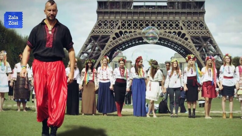 May include: A group of people wearing traditional Ukrainian clothing stand in front of the Eiffel Tower in Paris, France. The people are holding hands and smiling. A man in a black shirt and red pants walks towards the group.