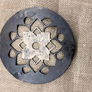 Könnte beinhalten: Ein runder, dunkelgrauer Metall-Topfuntersetzer mit floralem Design. Die Mitte zeigt eine silberfarbene, strukturierte Sternform. Der Untersetzer hat eine Reihe von ausgeschnittenen Formen am Rand und ist vor einem beigen, strukturierten Hintergrund platziert.
